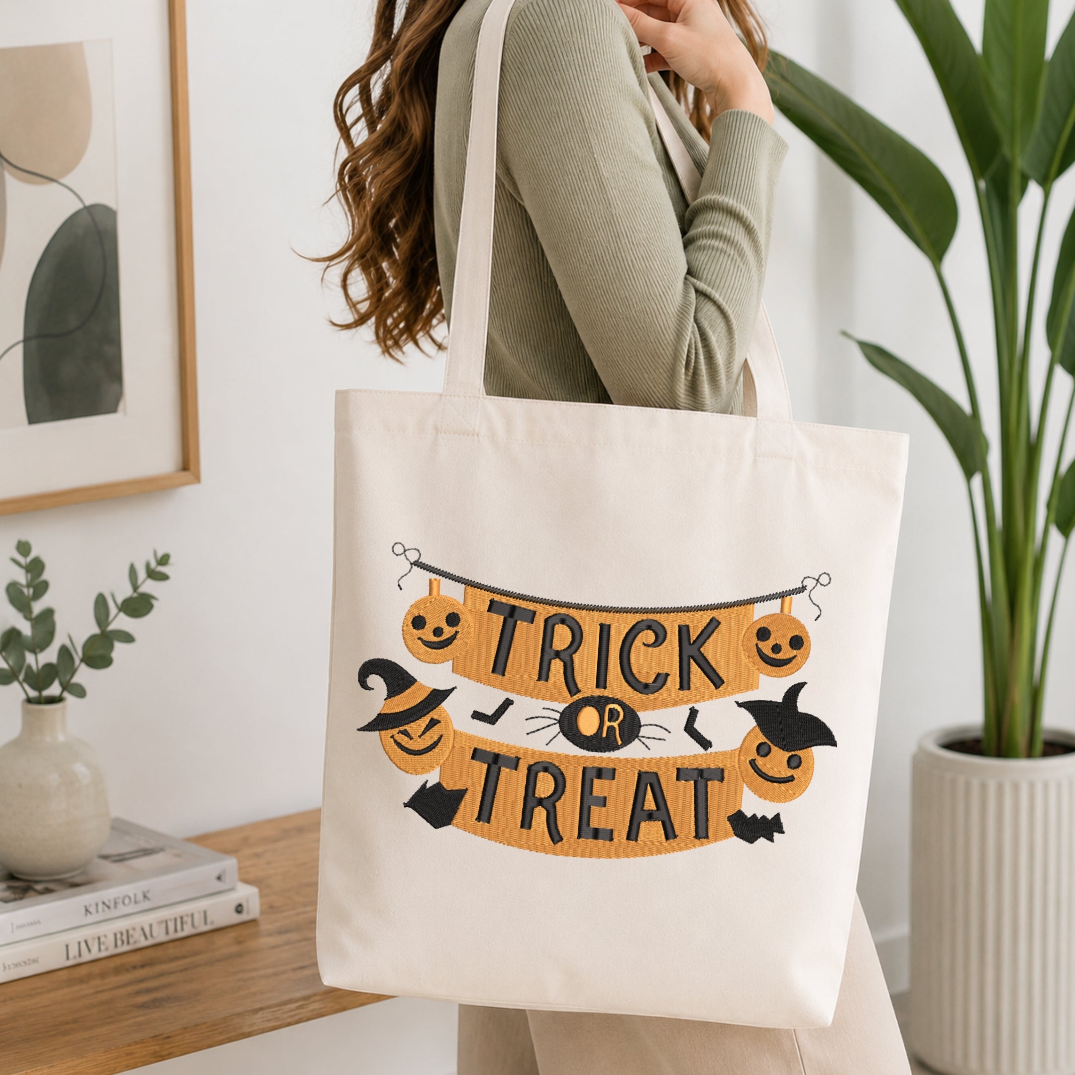 A woman wearing a sage green ribbed sweater carries a cream canvas tote bag with a Halloween graphic that reads 'TRICK OR TREAT' in black letters over an orange banner, decorated with jack-o'-lanterns, witch hats, bats, and a spider.