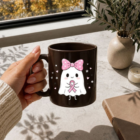 A hand holds a dark brown ceramic mug featuring a cute white ghost character with a pink bow and pink ribbon, set on a light stone tabletop near a window with a plant and candle in the background.
