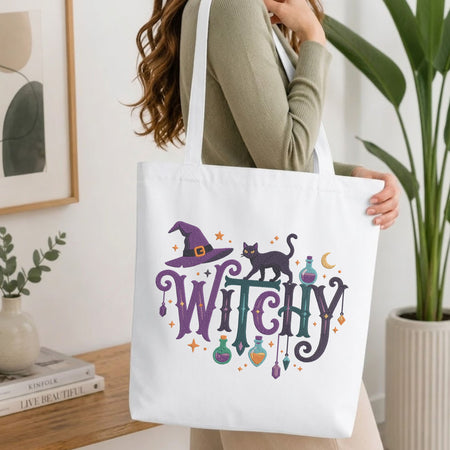 A woman wearing a sage green ribbed sweater carries a white canvas tote bag with a colorful 'Witchy' graphic featuring a purple witch hat, black cat, and potion bottles. The scene is set in a bright, modern room with a wooden table, framed art, and a potted plant.
