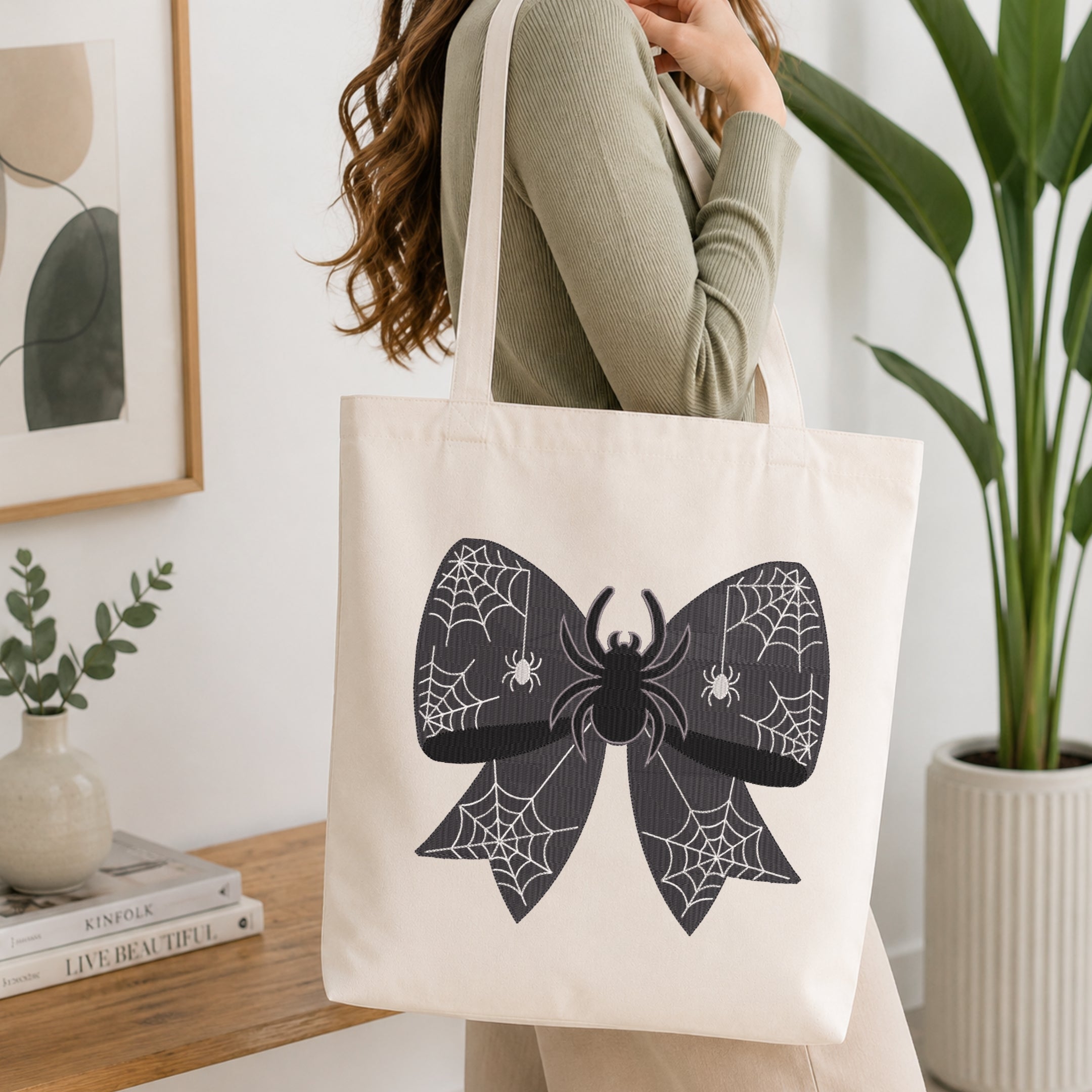 A person wearing a green long-sleeve top and carrying a natural canvas tote bag printed with the spider web bow machine embroidery graphic.