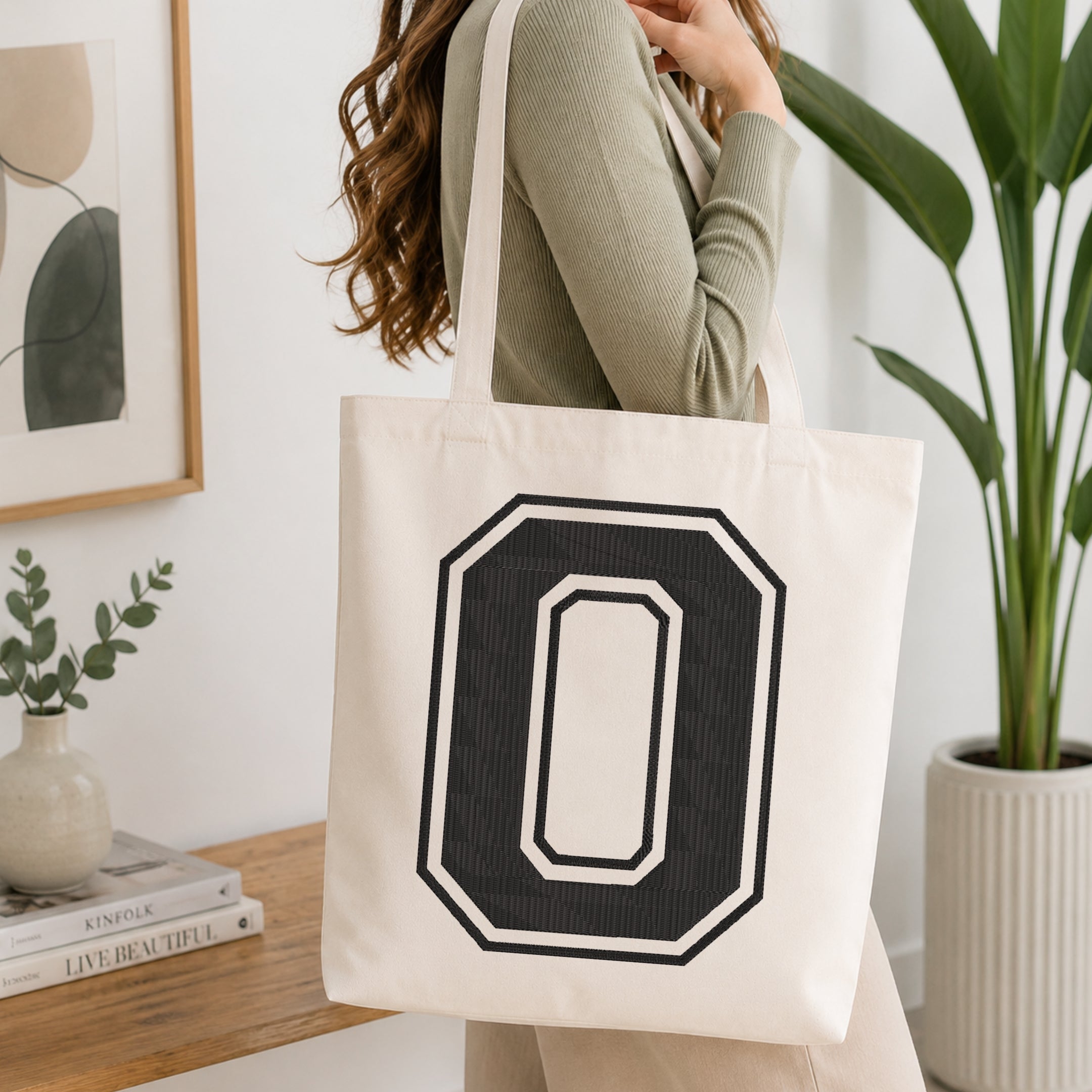 A woman wearing a sage green ribbed long-sleeve top carries a large off-white canvas tote bag with a bold black and white outlined letter 'O' on the front, set in a bright, modern room with a wooden console table, framed art, and a potted plant.
