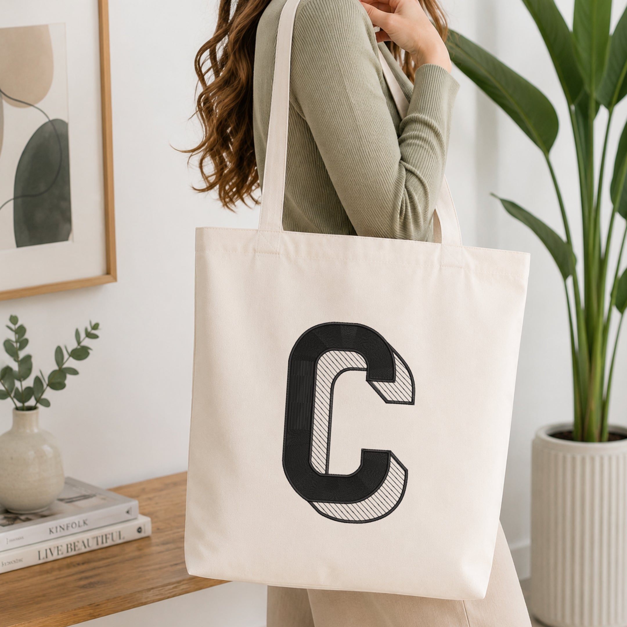 A woman wearing a sage green ribbed long-sleeve top carries a large off-white canvas tote bag with a bold black letter 'C' printed on the front, set in a bright, modern room with a wooden table, framed art, and a potted plant.