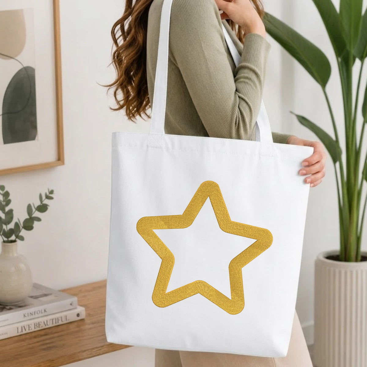 Woman carrying a white natural canvas tote bag featuring an embroidered golden star outline design in a bright, modern interior setting