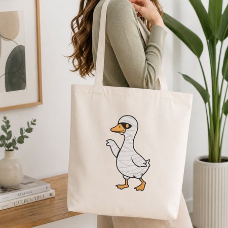 Woman carrying a white natural canvas tote bag featuring a cute duck in a mummy costume Halloween machine embroidery design in a bright, modern interior setting