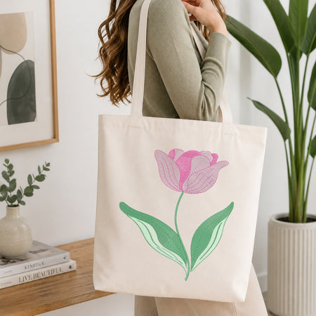 A woman wearing a sage green ribbed sweater carries a cream canvas tote bag with a pink tulip and green leaves printed on the front, set in a bright, modern room with a wooden table and potted plants.
