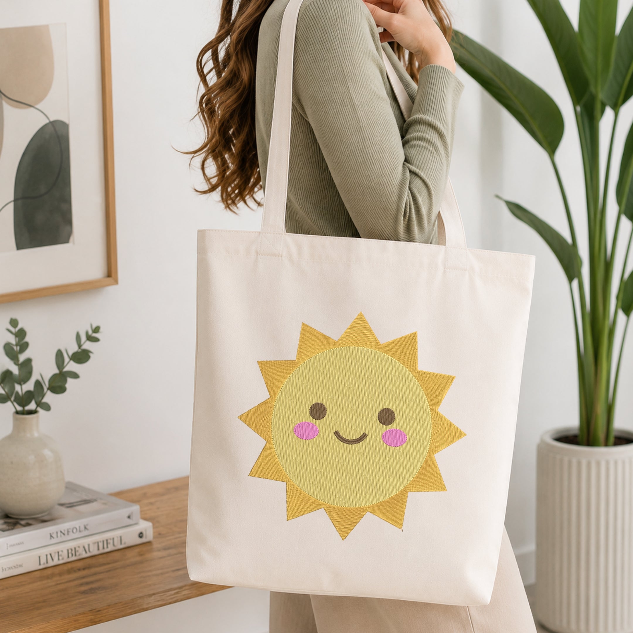 A woman wearing a sage green ribbed long-sleeve top carries a cream canvas tote bag with a cute yellow sun graphic on the front, set in a bright, modern room with a wooden table, books, and a potted plant.