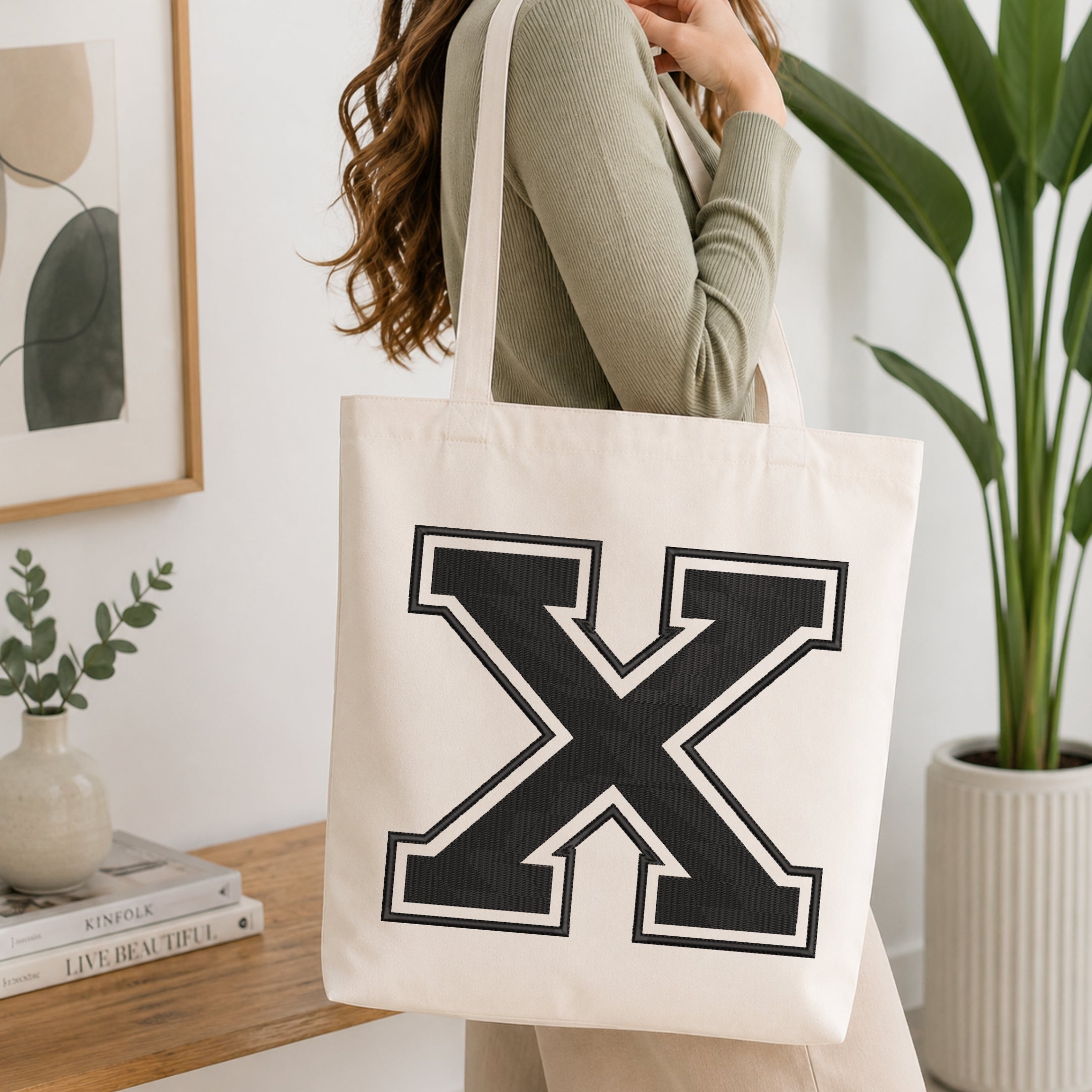 A person wearing a sage green ribbed top carries a large off-white canvas tote bag with a bold black block letter 'X' outlined in white on the front, set in a bright, modern room with a wooden console table and a potted plant.