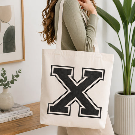 A person wearing a sage green ribbed top carries a large off-white canvas tote bag with a bold black block letter 'X' outlined in white on the front, set in a bright, modern room with a wooden console table and a potted plant.