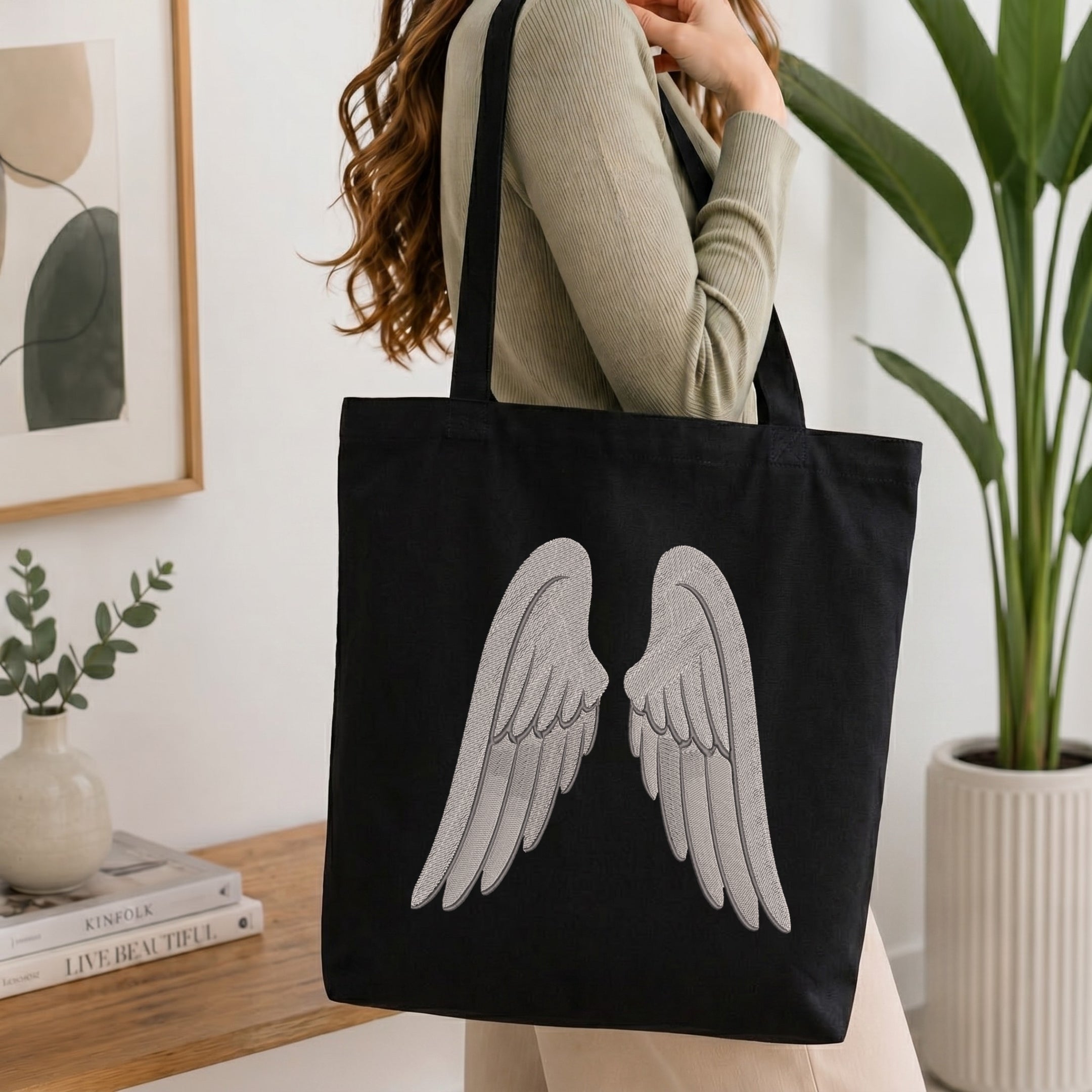 A woman wearing a beige ribbed sweater carries a black canvas tote bag with a large silver-gray embroidered angel wings design on the front, set against a bright, modern indoor background with a potted plant and framed art.