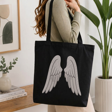 A woman wearing a beige ribbed sweater carries a black canvas tote bag with a large silver-gray embroidered angel wings design on the front, set against a bright, modern indoor background with a potted plant and framed art.