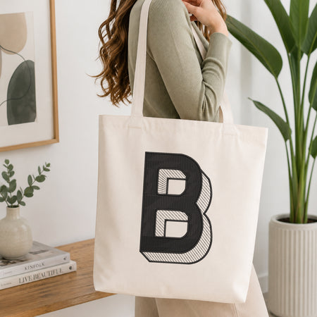 A woman wearing a sage green ribbed long-sleeve top carries a large off-white canvas tote bag with a bold black block letter 'B' printed on the front, set in a bright, modern room with a wooden table, framed art, and a potted plant.
