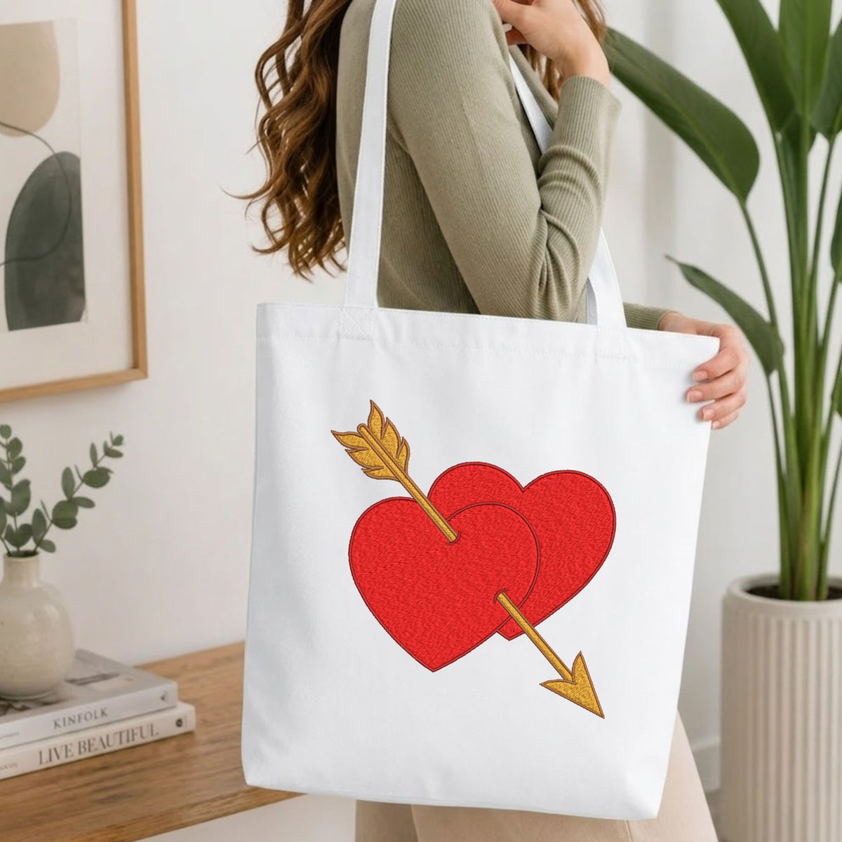 A woman wearing a sage green ribbed long-sleeve top carries a white canvas tote bag. The bag features a graphic of two red hearts overlaid with a gold arrow.