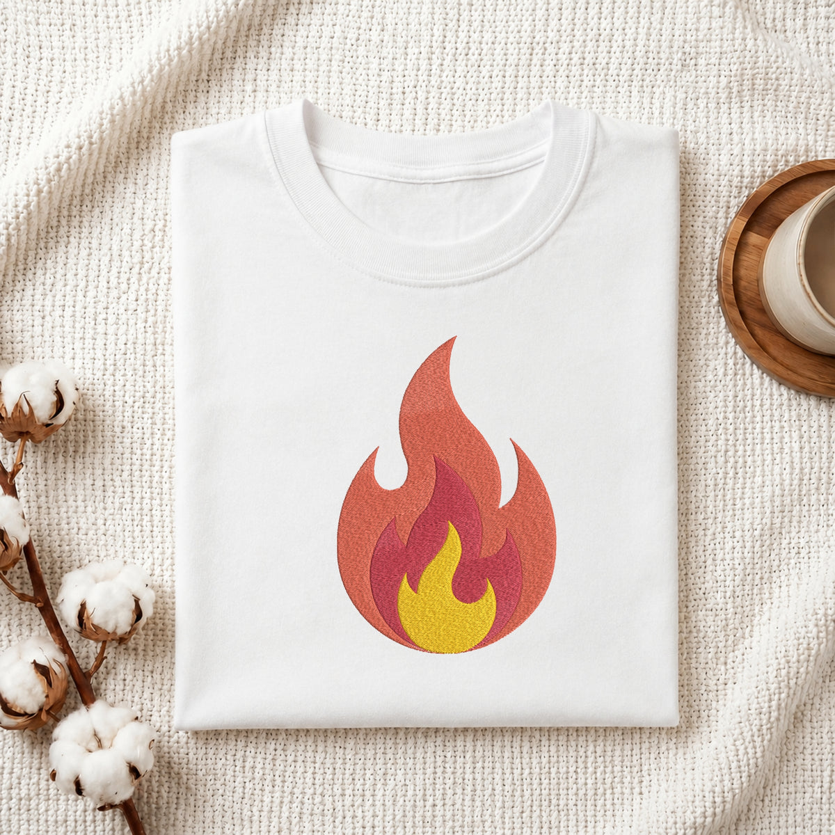 A folded white t-shirt with a stylized flame graphic in red, orange, and yellow, presented on a textured cream fabric background with cotton bolls and a wooden cup.