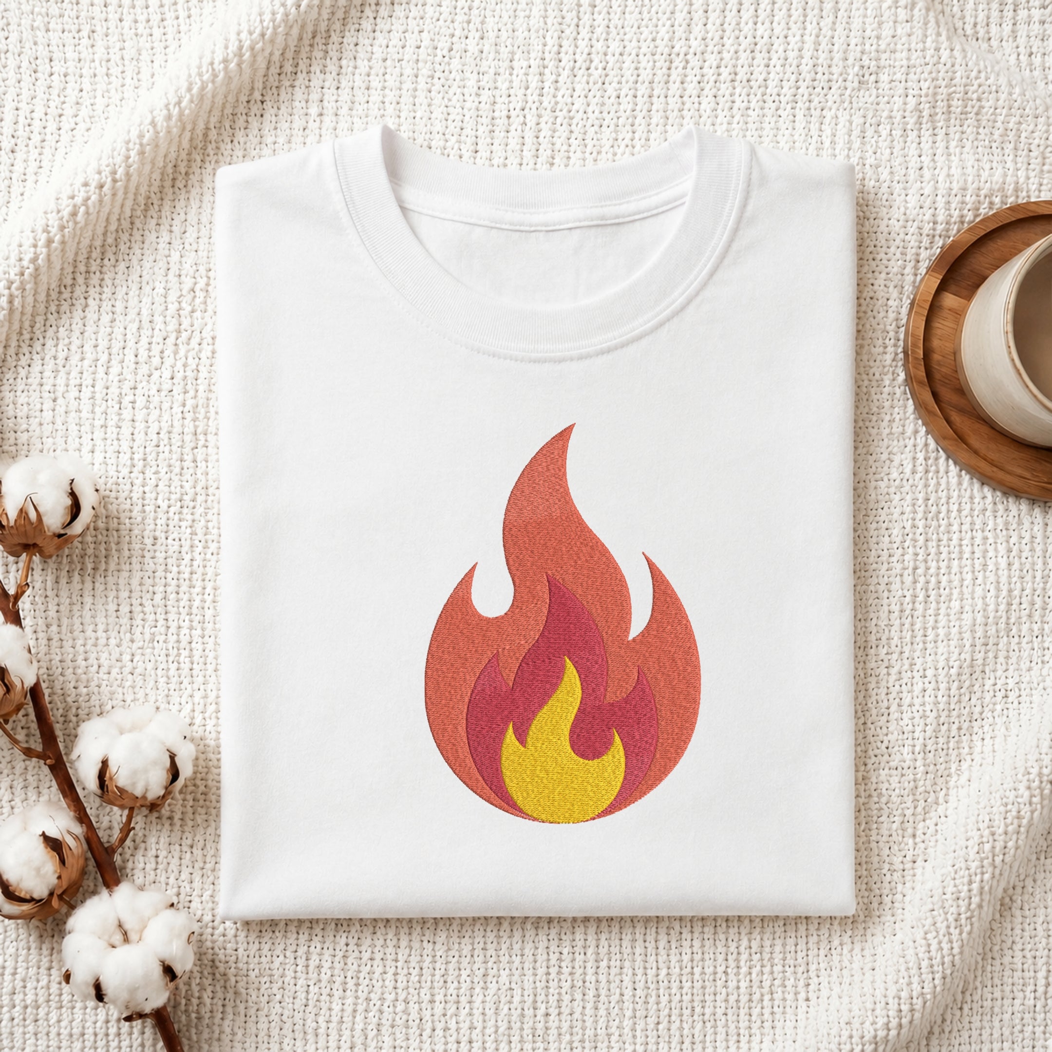 A folded white t-shirt with a stylized flame graphic in red, orange, and yellow, presented on a textured cream fabric background with cotton bolls and a wooden cup.