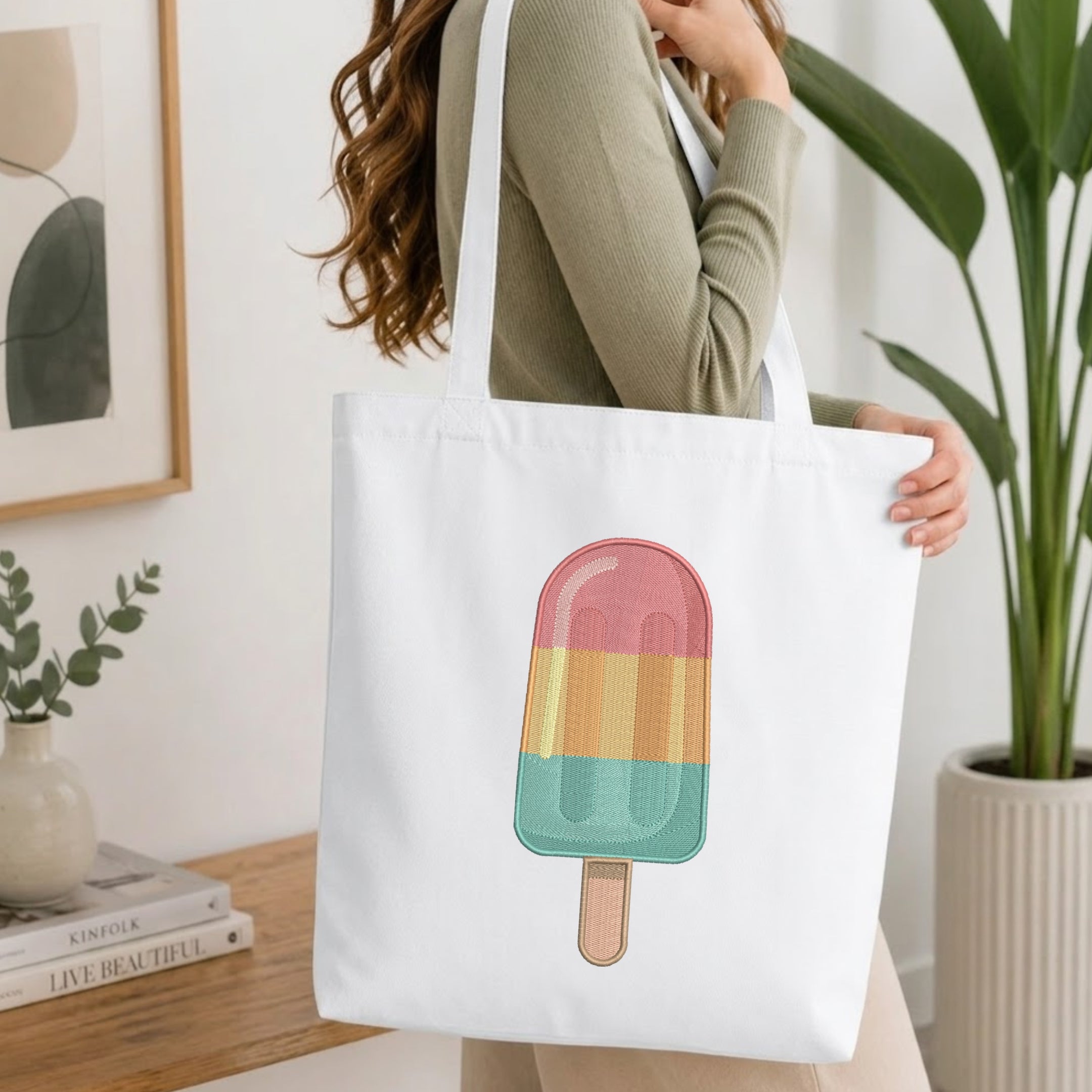 A woman wearing a sage green ribbed long-sleeve top carries a white canvas tote bag with a colorful popsicle graphic. The background is a bright, modern room with a wooden table, books, a framed print, and a potted plant.