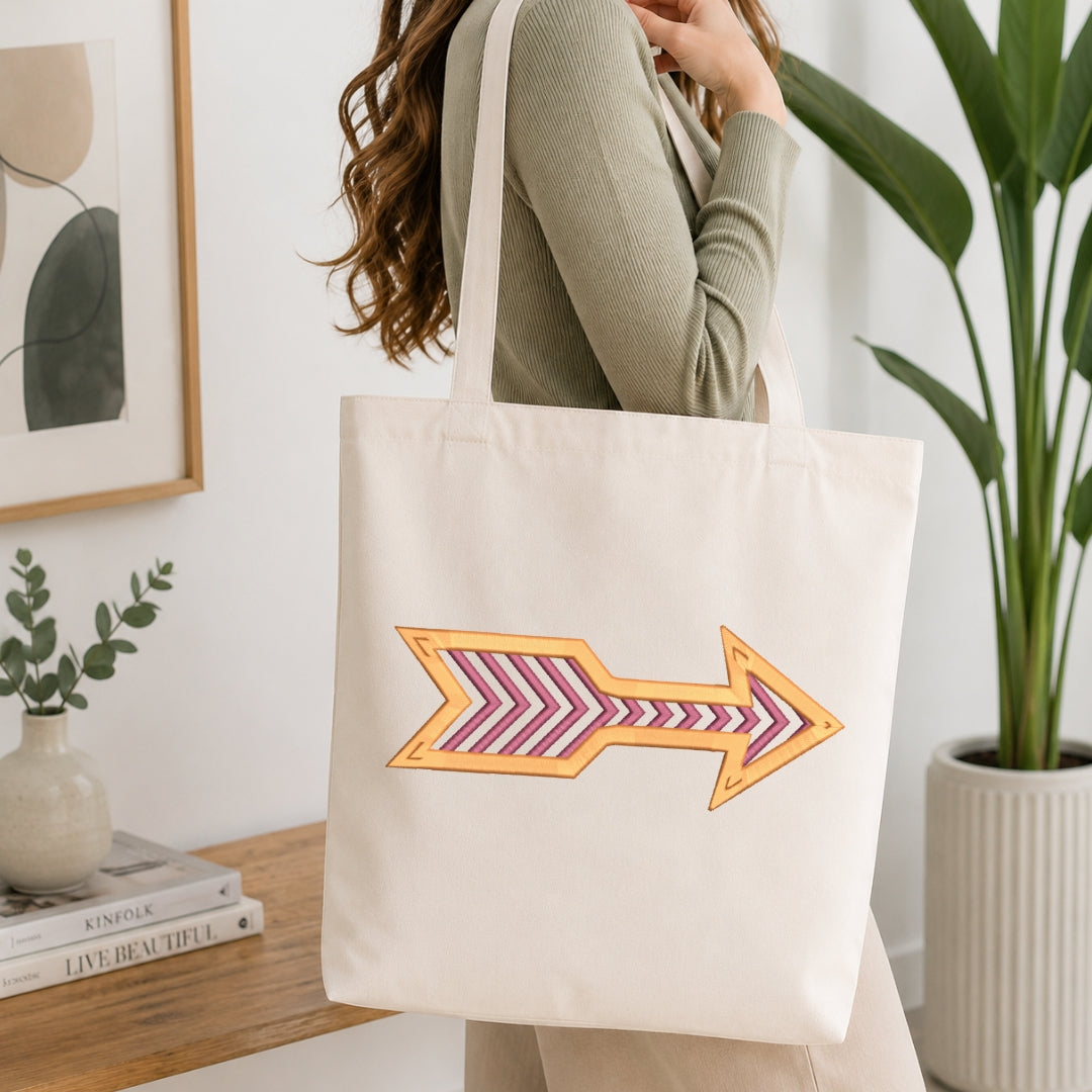 A woman wearing a sage green ribbed sweater carries a cream canvas tote bag with a large embroidered arrow design in orange and pink on the front, set in a bright, modern room with a wooden table and potted plants.