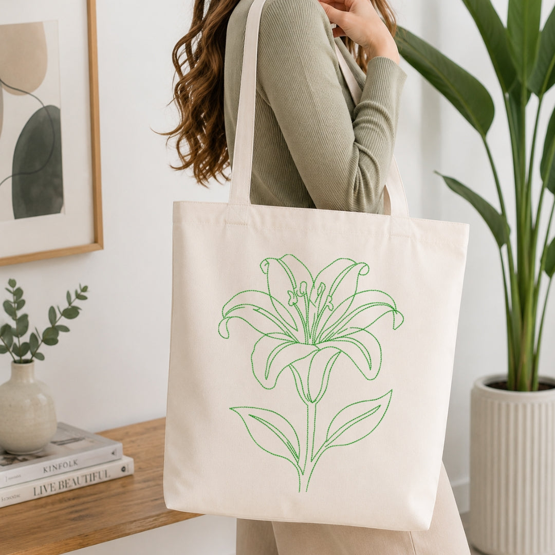 A person wearing a ribbed sage green long-sleeve top carries a large off-white canvas tote bag with a green line-art lily design. The scene is set in a bright, modern room with a wooden table, books, a small vase, and a potted plant.