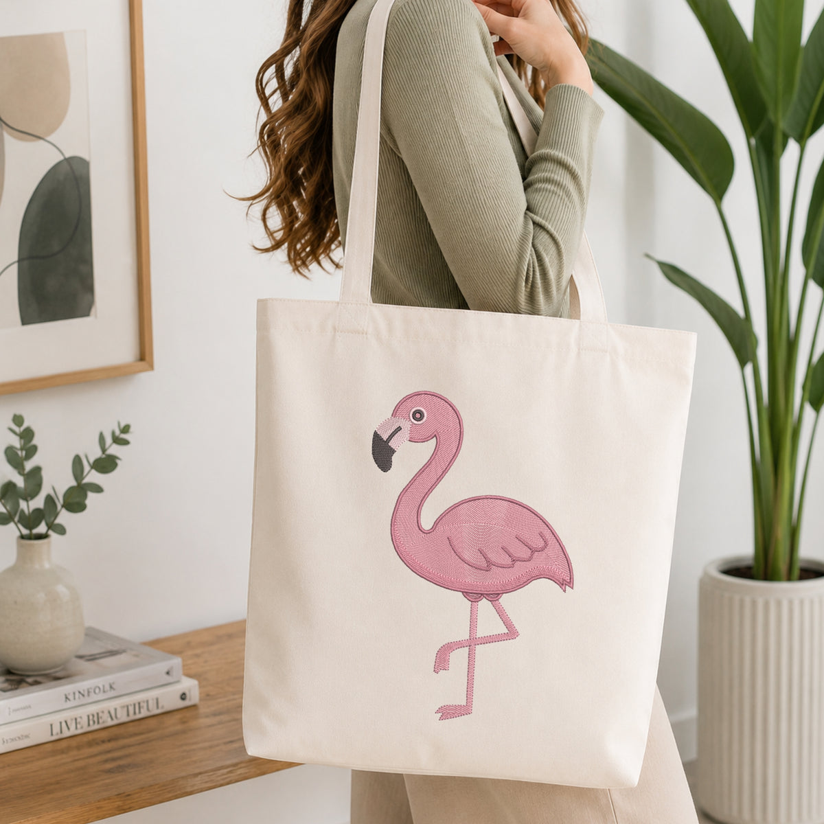 A woman wearing a sage green ribbed sweater carries a cream canvas tote bag with a pink flamingo graphic printed on the front, set in a bright, modern room with a wooden table, framed art, and a potted plant.