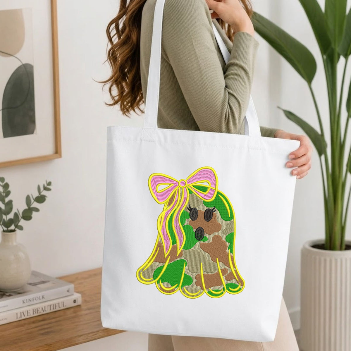 Woman carrying a white natural canvas tote bag featuring an embroidered camo ghost design in a bright, modern interior setting