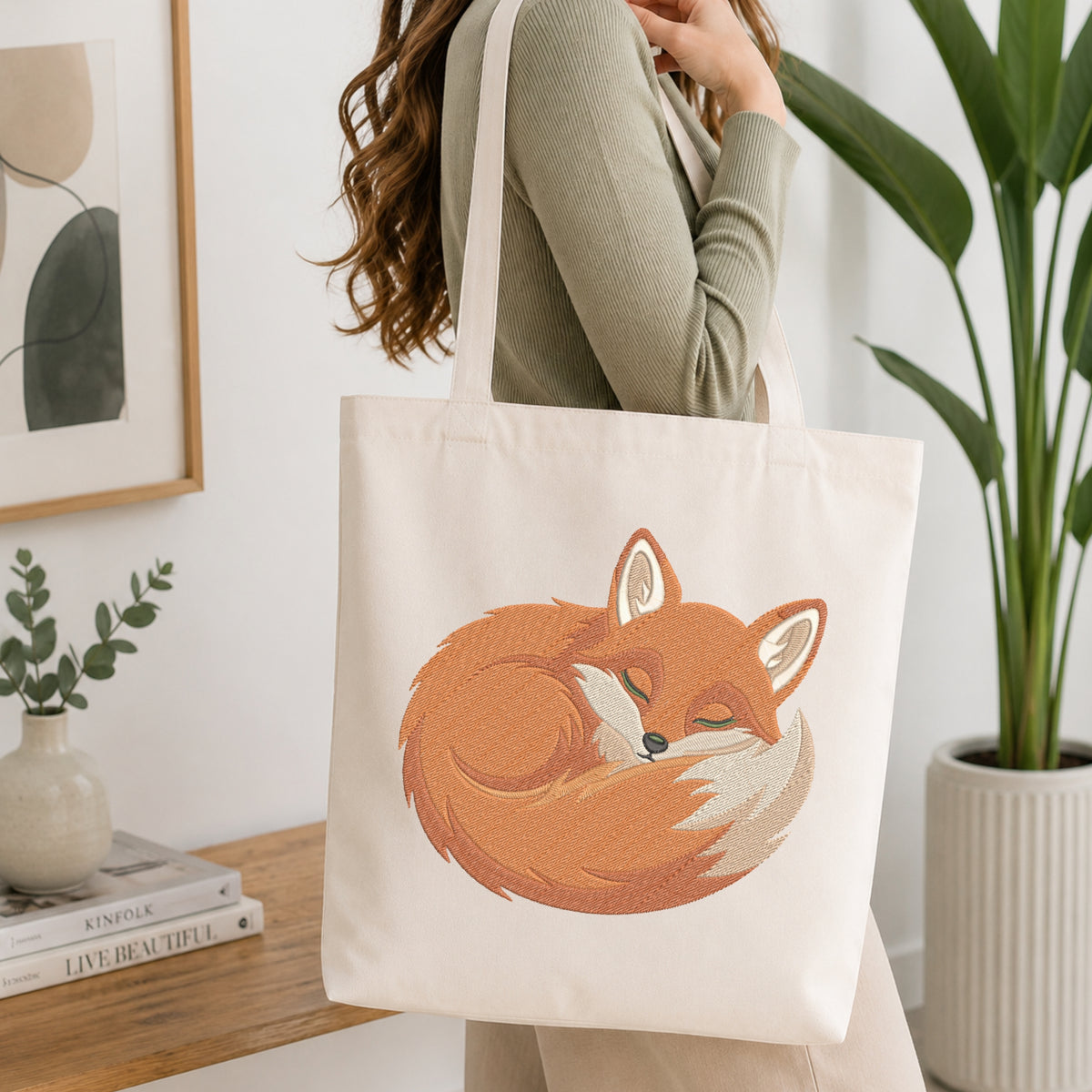 A woman wearing a sage green ribbed sweater carries a cream canvas tote bag with a large embroidered sleeping fox design in orange, brown, and white. The scene is set in a bright, modern room with a wooden table, books, a framed print, and a potted plant.