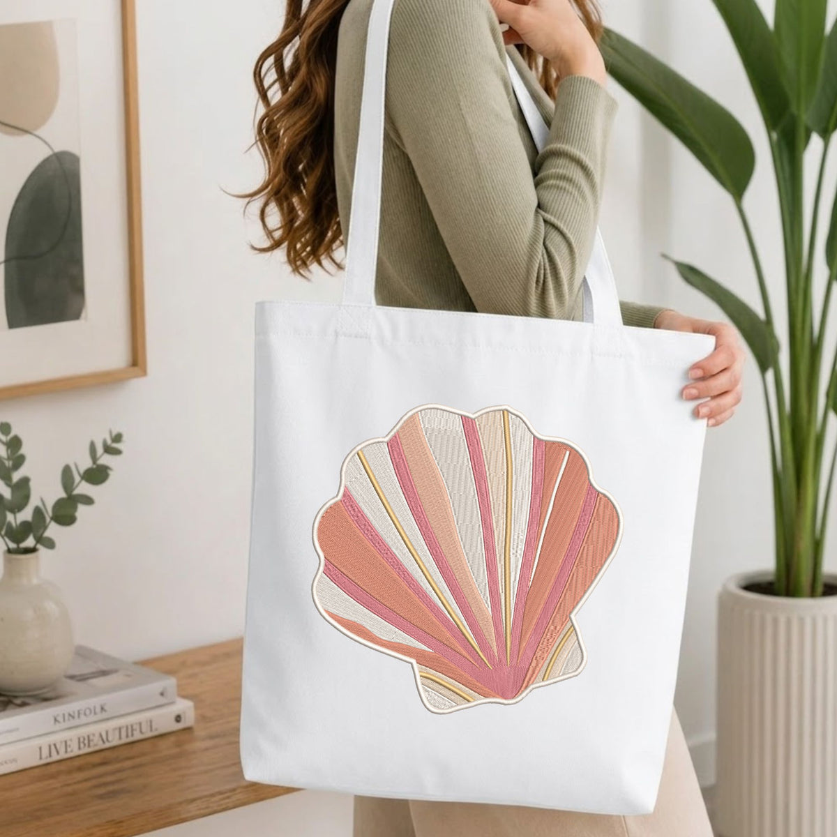 Woman carrying a white natural canvas tote bag featuring an embroidered pink and beige coastal seashell design in a bright, modern interior setting.