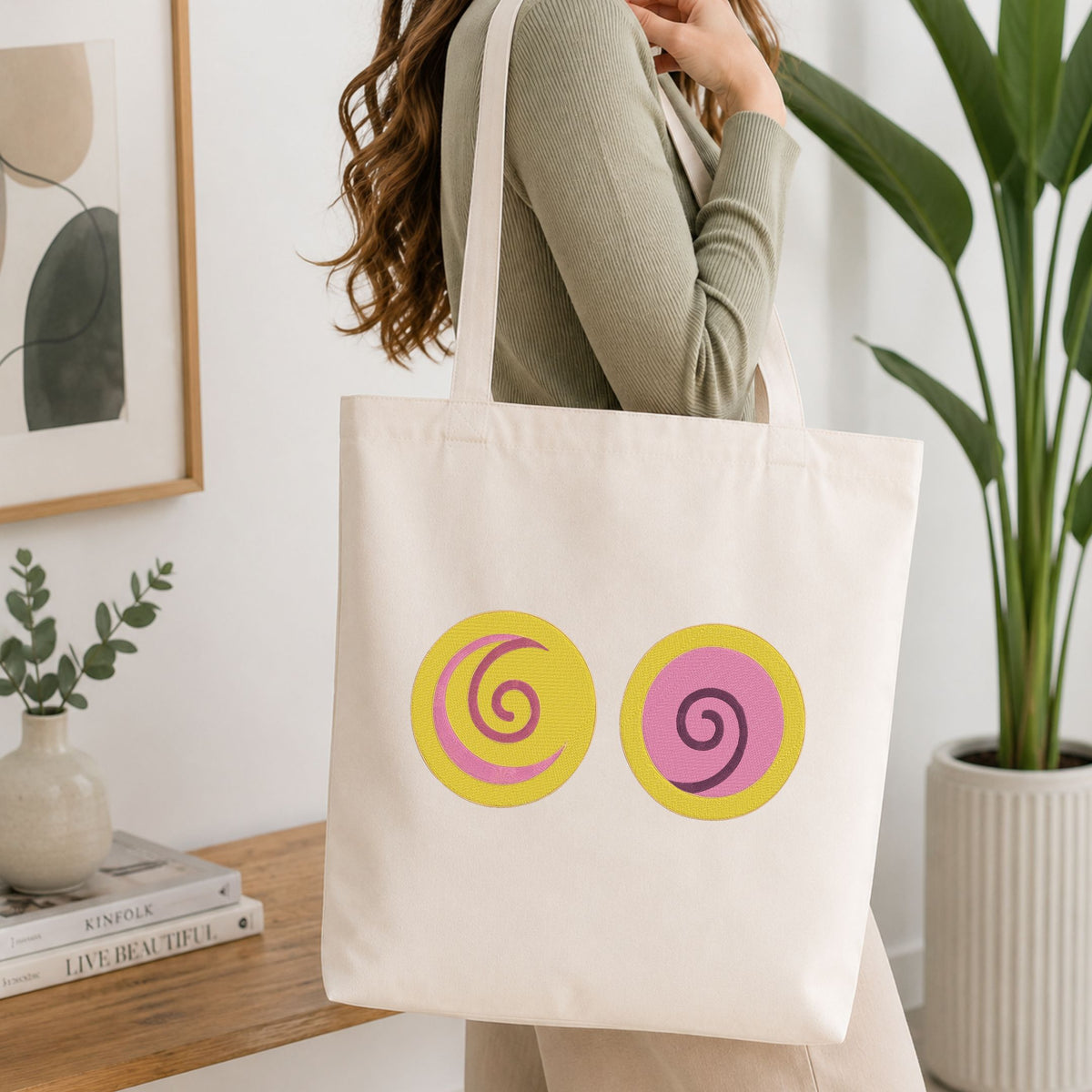 A woman wearing a sage green ribbed sweater carries a cream canvas tote bag with two yellow circular graphics featuring pink and purple spirals, set in a bright, modern room with a wooden table and potted plants.
