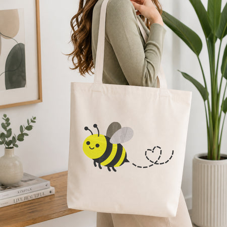 A woman wearing a sage green ribbed sweater carries a cream canvas tote bag with a cute yellow and black bumblebee graphic and a dashed heart tail. The scene is set in a bright, modern room with a wooden table, books, a small plant, and a large green potted plant in the background.