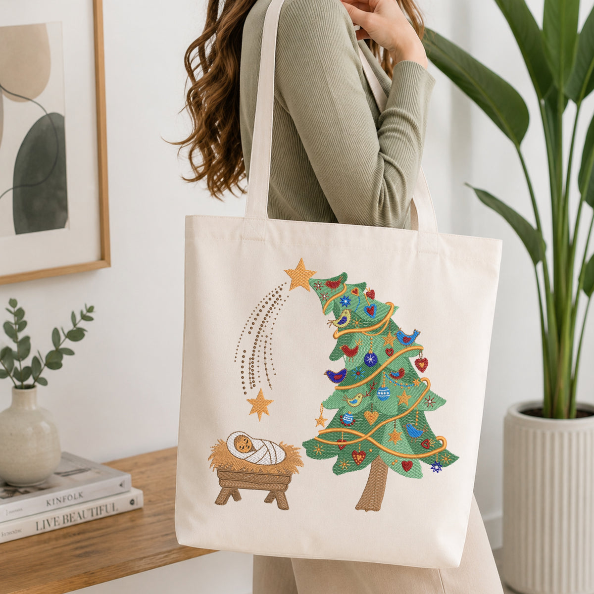 A woman wearing a sage green ribbed long-sleeve top carries a cream canvas tote bag with a colorful embroidered Christmas tree, star, and baby nativity scene on the front, set against a bright, modern interior background.