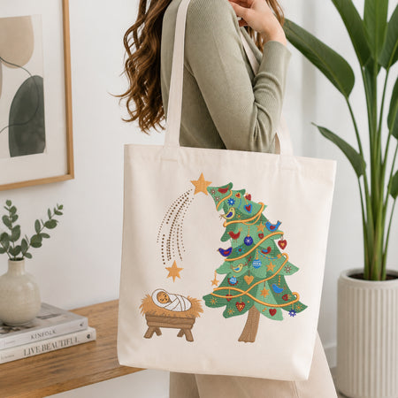 A woman wearing a sage green ribbed long-sleeve top carries a cream canvas tote bag with a colorful embroidered Christmas tree, star, and baby nativity scene on the front, set against a bright, modern interior background.