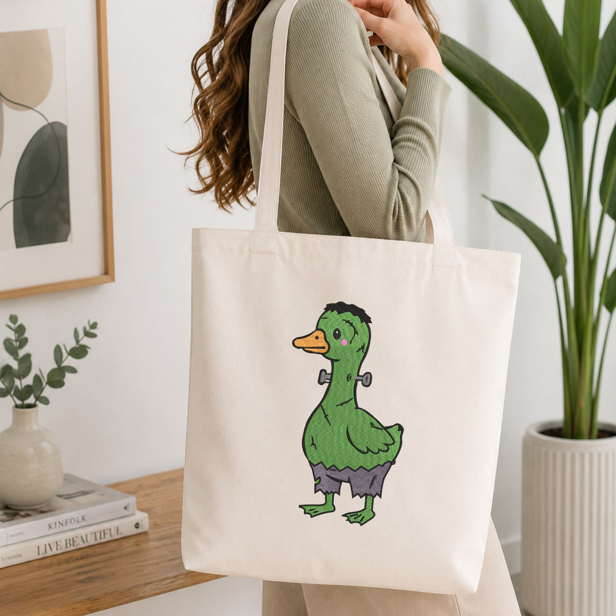 Woman carrying a white natural canvas tote bag featuring a cute green Frankenstein monster duck Halloween machine embroidery design in a bright, modern interior setting
