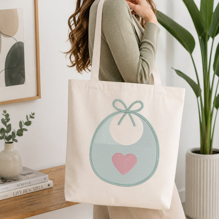 A woman wearing a sage green ribbed long-sleeve top carries a cream canvas tote bag with a graphic of a light blue bib, white circle, and pink heart. The scene is set in a bright, modern room with a wooden table, books, a small plant, and a large green potted plant.