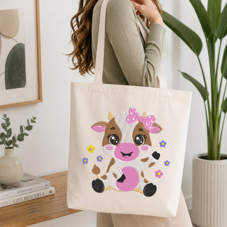A person holding a natural canvas tote bag printed with an embroidered cute sitting baby cow illustration, set against a bright interior room background with green plants.