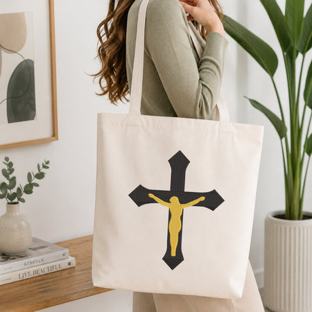 Woman carrying a beige canvas tote bag customized with a black and gold crucifix embroidery design.