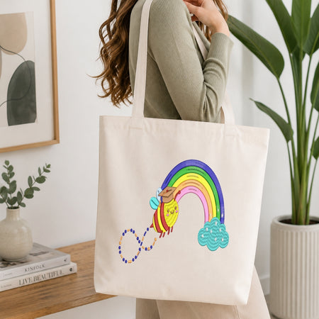A woman wearing a ribbed sage green long-sleeve top carries a cream canvas tote bag. The bag features a colorful embroidered design of a rainbow arching over a yellow sun and a blue cloud, with a beaded line connecting them.