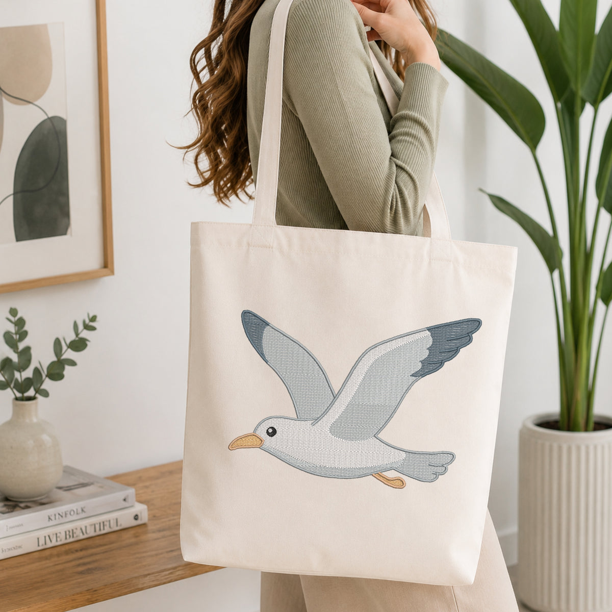 A woman wearing a sage green ribbed sweater carries a cream canvas tote bag with a detailed embroidered flying seagull design. The setting is a bright, modern room with a wooden shelf, framed art, and a potted green plant.