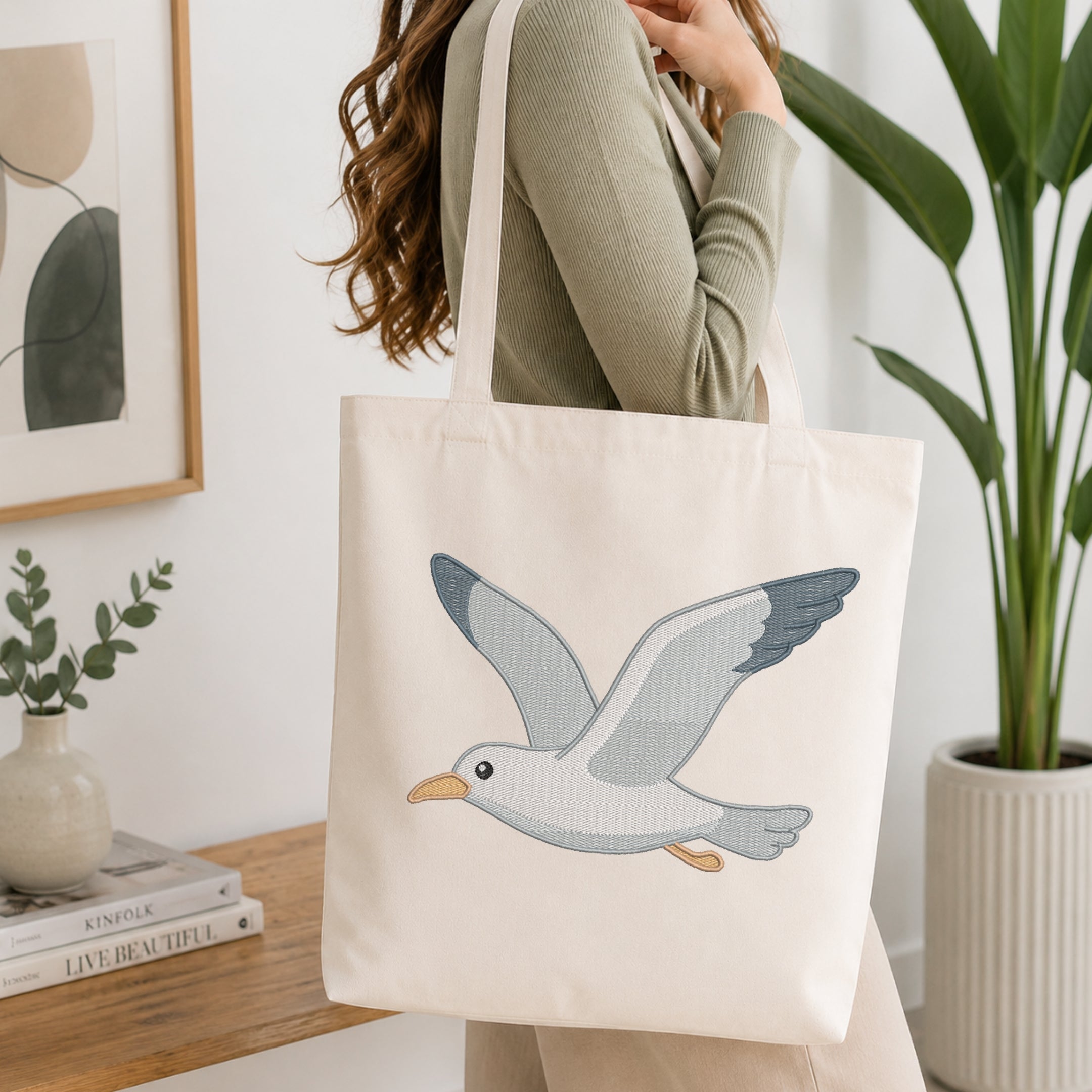 A woman wearing a sage green ribbed sweater carries a cream canvas tote bag with a detailed embroidered flying seagull design. The setting is a bright, modern room with a wooden shelf, framed art, and a potted green plant.