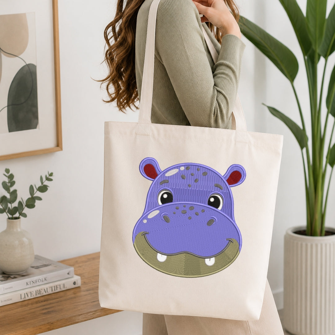 A woman wearing a ribbed sage green long-sleeve top carries a cream canvas tote bag with a large purple and green hippo face graphic printed on the front, set in a bright, modern room with a wooden table and potted plants.