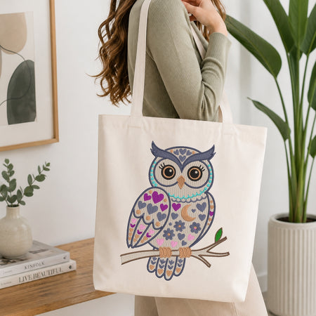 A woman holds a cream canvas tote bag featuring a colorful embroidered owl design with hearts, crescent moons, and floral patterns on a branch. The setting is a bright, modern room with a wooden table, books, a potted plant, and framed wall art.