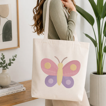A woman wearing a sage green ribbed sweater carries a cream canvas tote bag with a colorful butterfly design featuring pink, purple, and yellow wings. The scene is set in a bright, modern room with a wooden table, books, a small plant, and a large green potted plant in the background.