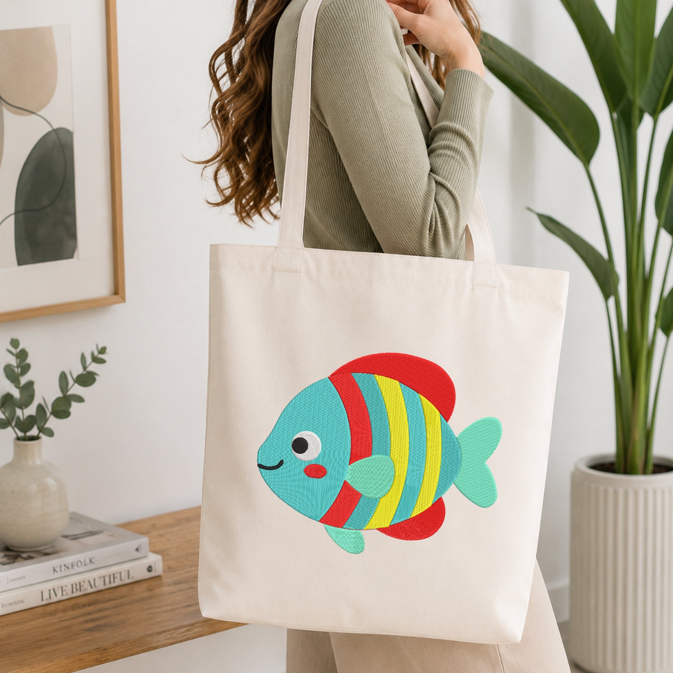 A woman wearing a sage green ribbed sweater carries a cream canvas tote bag with a colorful embroidered tropical fish. The fish features red, yellow, and blue stripes with a light blue-green body and tail.