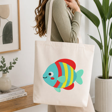 A woman wearing a sage green ribbed sweater carries a cream canvas tote bag with a colorful embroidered tropical fish. The fish features red, yellow, and blue stripes with a light blue-green body and tail.