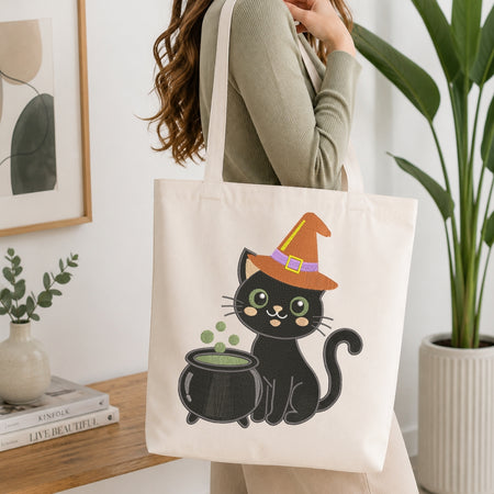 A woman wearing a sage green ribbed sweater carries a cream canvas tote bag featuring a cartoon black cat in an orange witch hat next to a cauldron. The scene is set in a bright, modern room with a wooden table, books, a small plant, and a large green potted plant in the background.