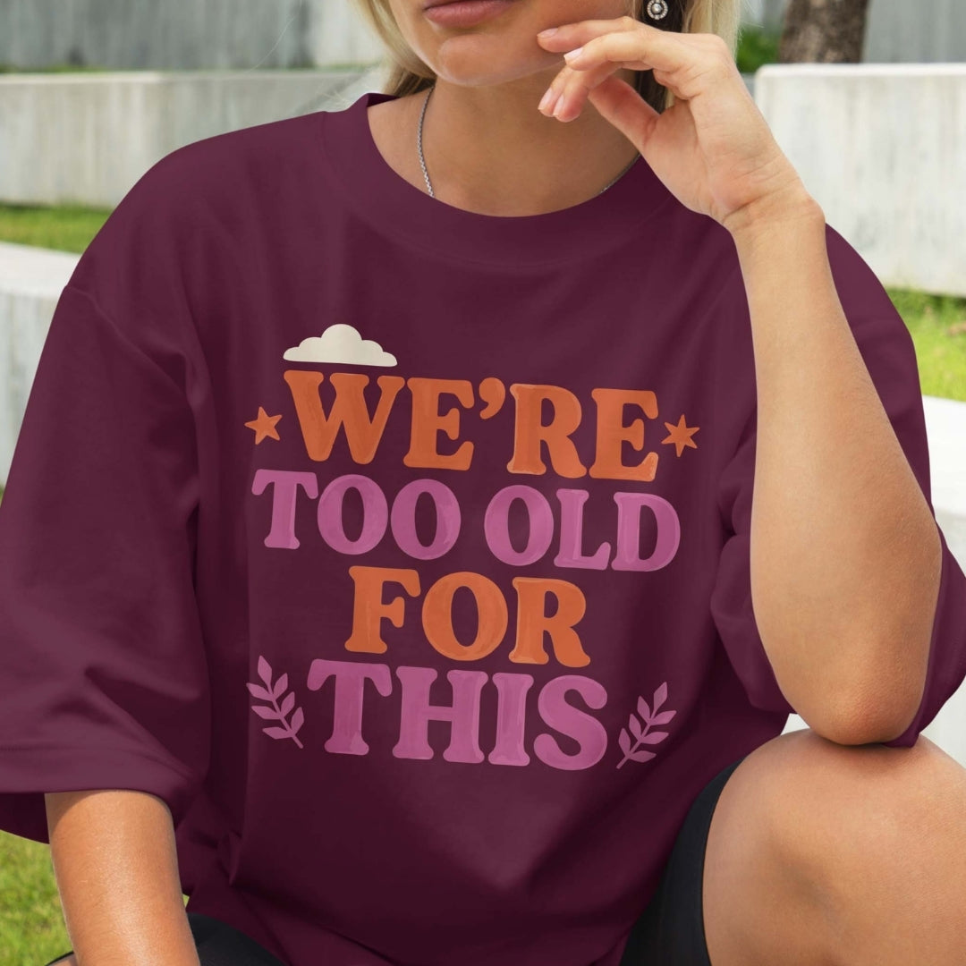 Person wearing a purple t-shirt with text 'WE'RE TOO OLD FOR THIS' in an outdoor setting.