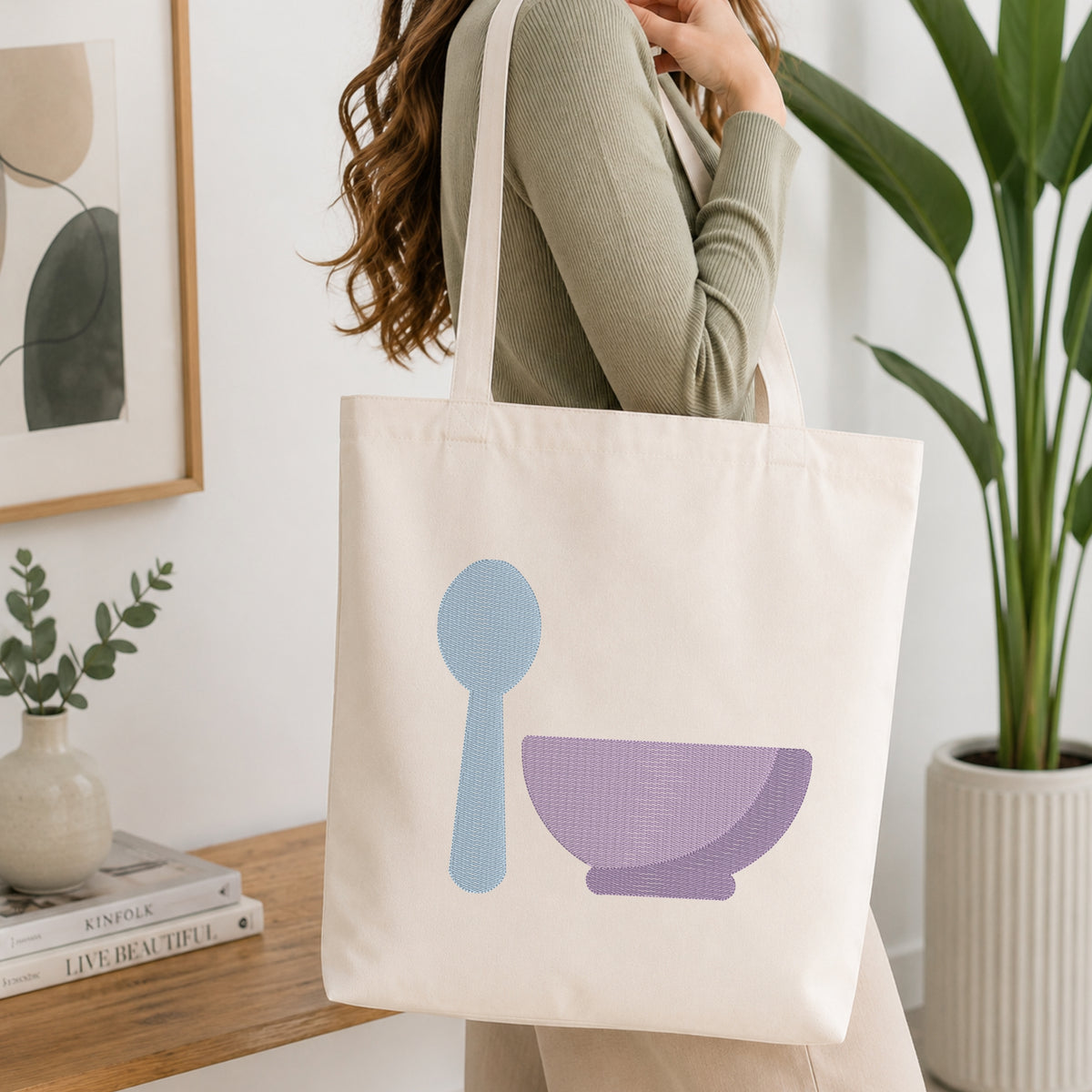 A woman wearing a sage green ribbed sweater carries a cream canvas tote bag with a light blue spoon and purple bowl graphic, set in a bright, modern room with a wooden table, books, and a potted plant.
