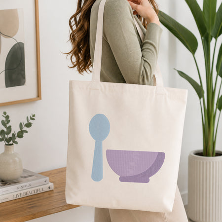 A woman wearing a sage green ribbed sweater carries a cream canvas tote bag with a light blue spoon and purple bowl graphic, set in a bright, modern room with a wooden table, books, and a potted plant.