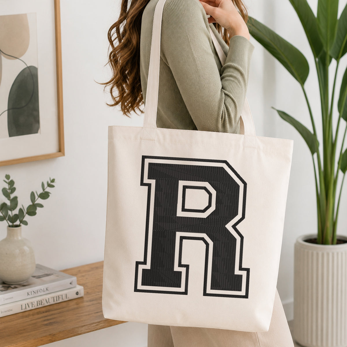 A person carrying a white canvas tote bag featuring a large embroidered varsity block letter 'R' in black with a white outline, set in a bright room with green house plants.