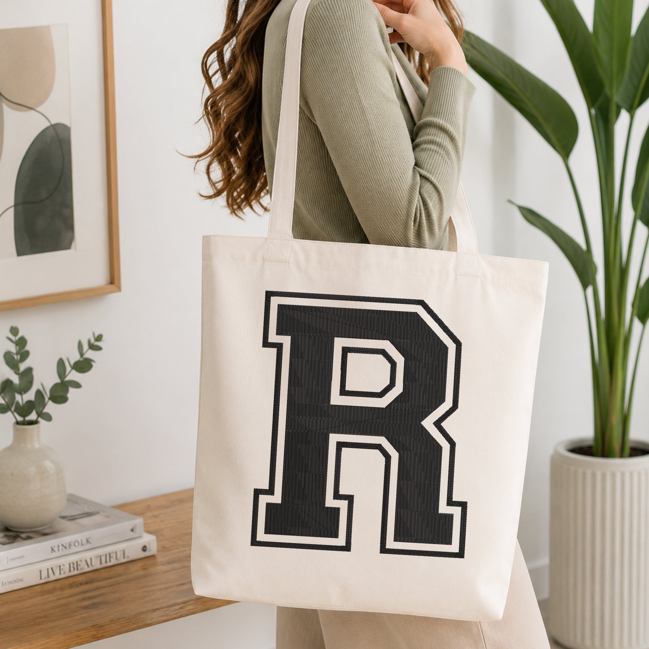 A person carrying a white canvas tote bag featuring a large embroidered varsity block letter 'R' in black with a white outline, set in a bright room with green house plants.
