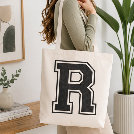 A person carrying a white canvas tote bag featuring a large embroidered varsity block letter 'R' in black with a white outline, set in a bright room with green house plants.