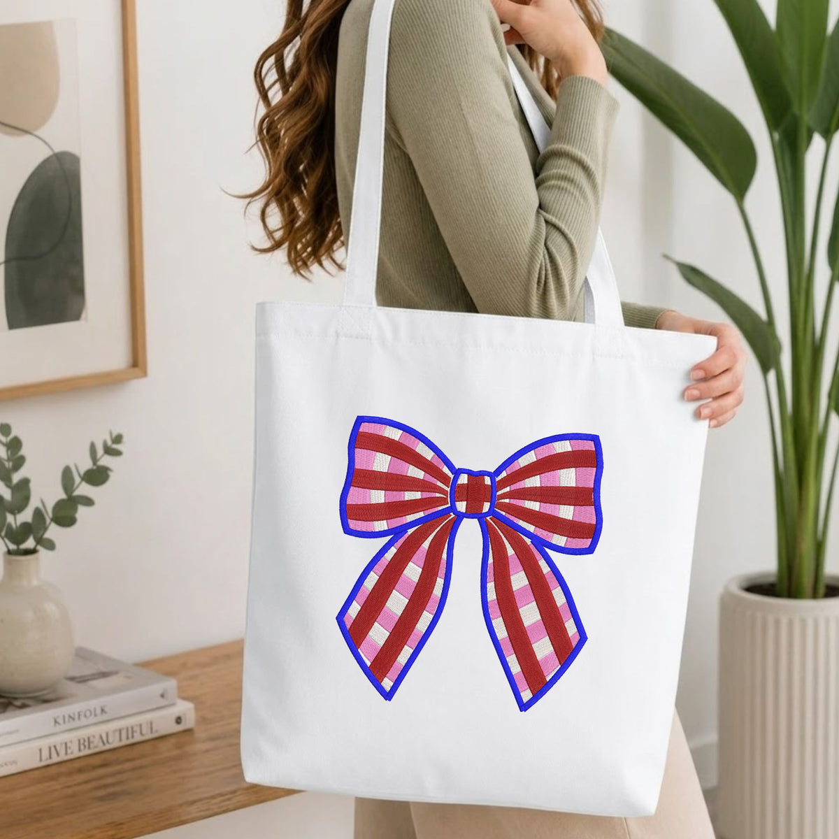 Woman carrying a white natural canvas tote bag featuring an embroidered red and pink plaid bow design in a bright, modern interior setting.