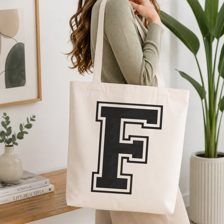 A woman wearing a sage green ribbed sweater carries a large off-white canvas tote bag with a bold black block letter 'F' outlined in white on the front, set in a bright, modern room with a wooden console table and a potted plant.